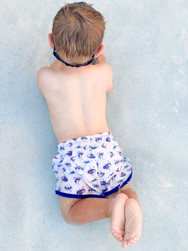 a young boy in swimming trunks laying on his stomach on a sandy beach.