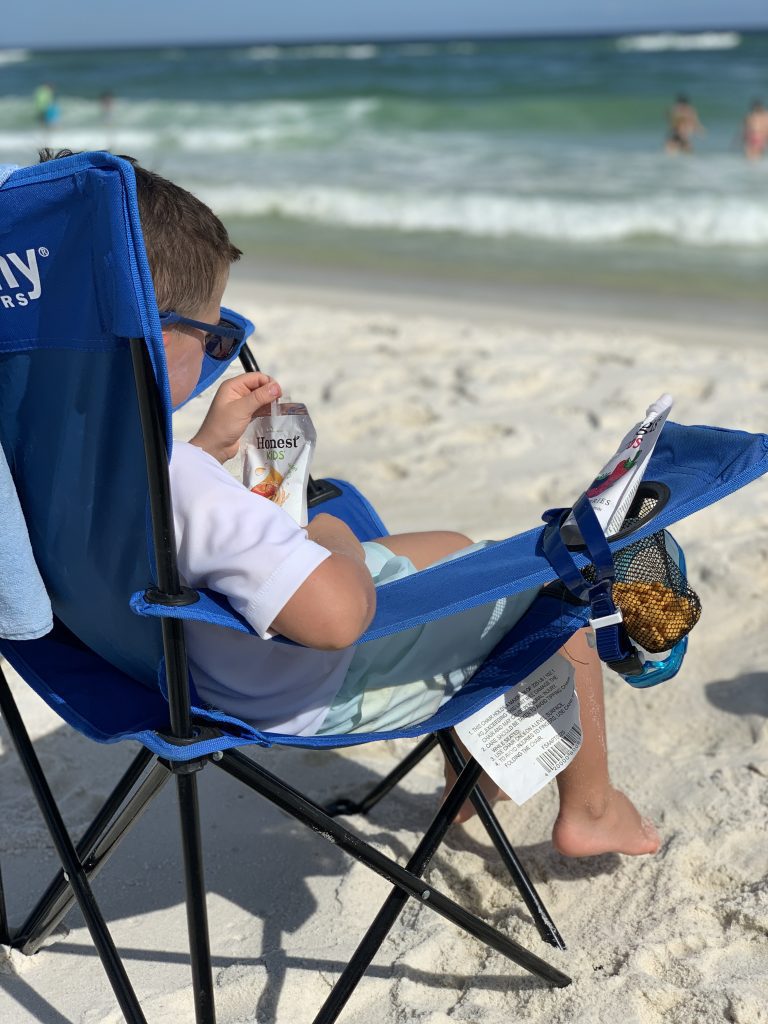 a little boy lounging in a beach chair drinking a juice box and having a snack.