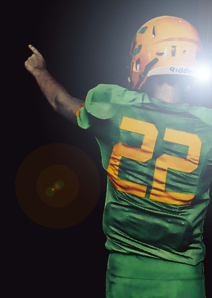 The back of a tackle football player in a green uniform with yellow 22 and a yellow helmet. He is pointing to the sky with his left hand.