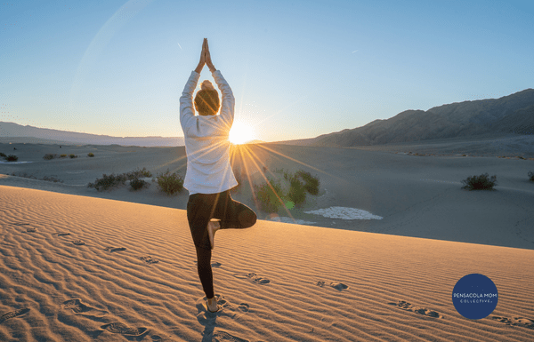 Woman doing yoga tree pose on sand with sunlight shining through.