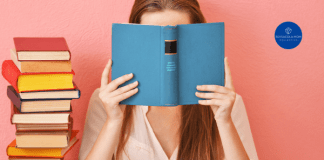 a woman holding a book with a blue cover in front of her face. there is a tall stack of books next to her.