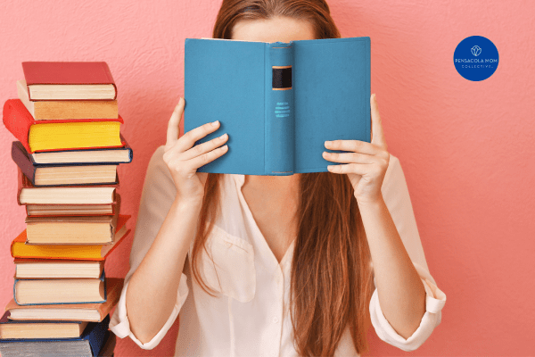 a woman holding a book with a blue cover in front of her face. there is a tall stack of books next to her.