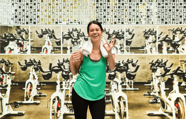 woman smiling in front of bicycles in a cycling studio. She has a towel around her neck, holding a bottle of water in her right hand and giving the ok sign with her left.