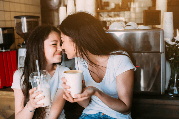 Mom and daughter touching noses. Mom is holding a coffee mug and daughter is holding a smoothie