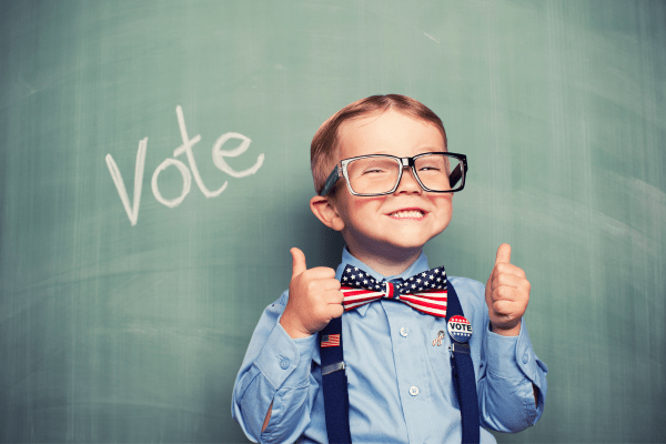 Little boy with glasses, blue shirt, suspenders and a bow tie with American flag print standing in front of a chalk board with the word "vote" written on it.