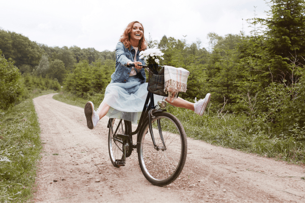 Woman in a dress and jean jacket riding a bike down a dirt road with her feet in the air