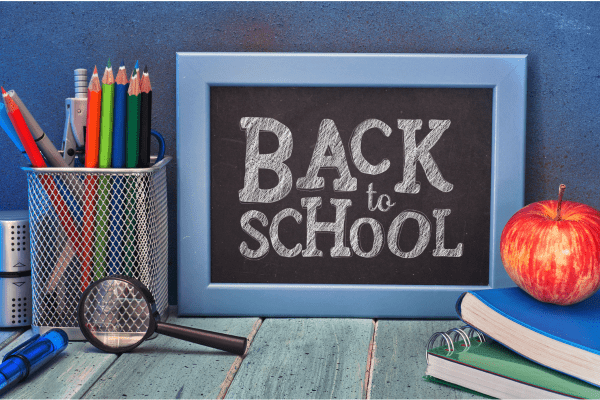 a container of pencils, a magnifying glass, a stack of two notebooks with an apple on top and a chalkboard sign that says back to school