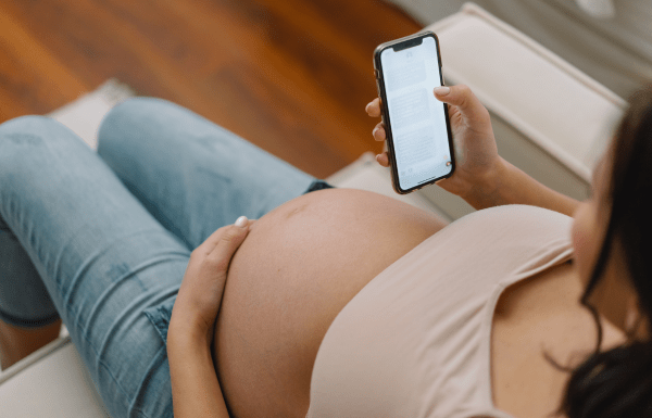 An overhead photo of a pregnant woman scrolling on her phone. Her bare belly is exposed and she's wearing jeans.