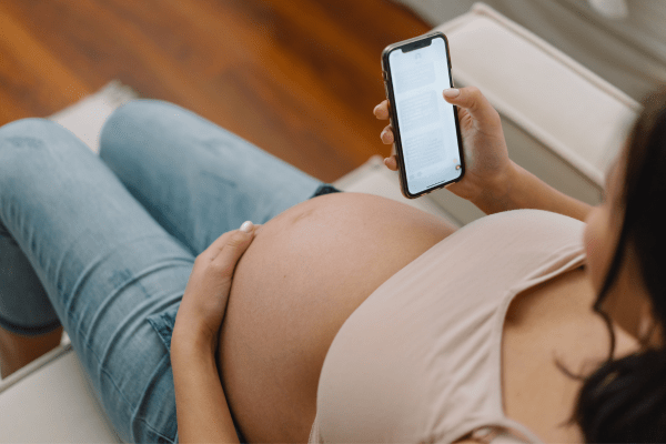 An overhead photo of a pregnant woman scrolling on her phone. Her bare belly is exposed and she's wearing jeans.