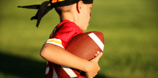 A stock photo of a young boy playing flag football. He is carrying the football and running with it. He is wearing a red jersey and a black headband.