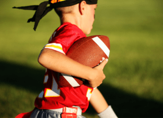 A stock photo of a young boy playing flag football. He is carrying the football and running with it. He is wearing a red jersey and a black headband.