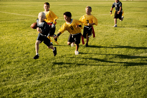 Five young boys playing flag football on a grass field. One boy is pulling the flag of the child carrying the football.