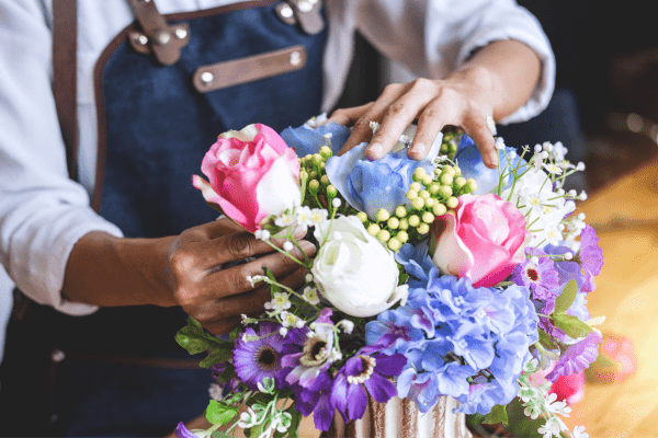 An African American woman wearing a dark blue apron, her face is not pictured but she is a florist creating a fresh flower arrangement.
