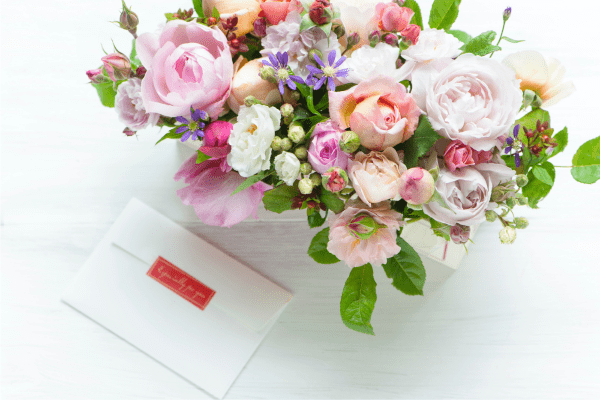 A fresh flower arrangement photographed from above. The flowers are light pink, lavender and white with greenery. and And there is an envelope sitting next to the arrangement.