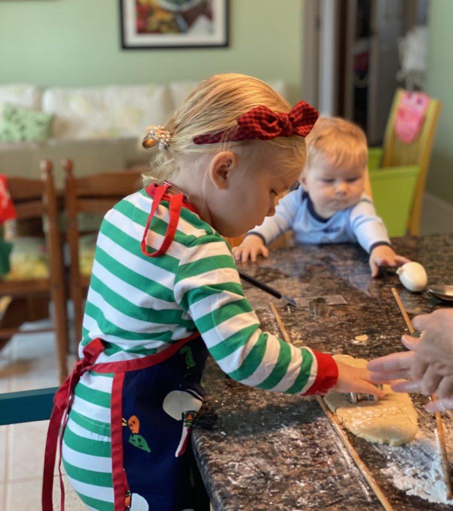A toddler girl helping in the kitchen with her baby brother sitting next to her.