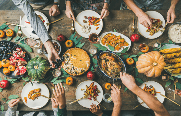 An overhead shot of a dining table set for Thanksgiving with 6 sets of hands eating and serving from the dishes of turkey, squash, cranberries and potatoes.
