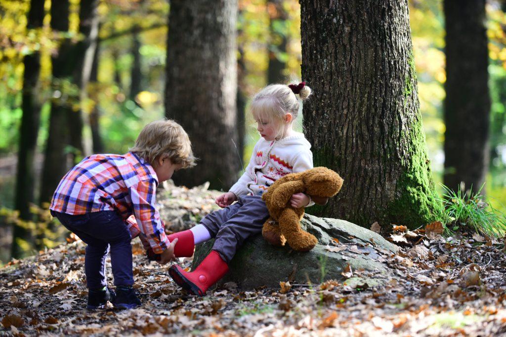 One child helping another put her boot on in the woods.