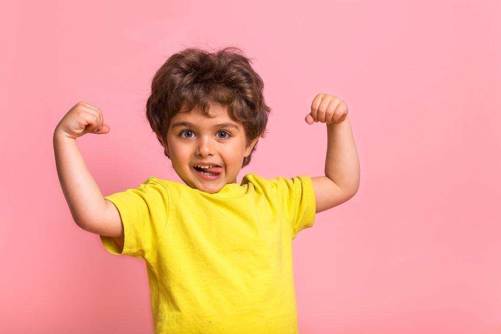 Funny little sportive kid boy in yellow shirt, showing his muscles. Happy strong nerd kindergarten child showing bicep. Dream, confidence, success, possible, innovation. Go back elementary school 