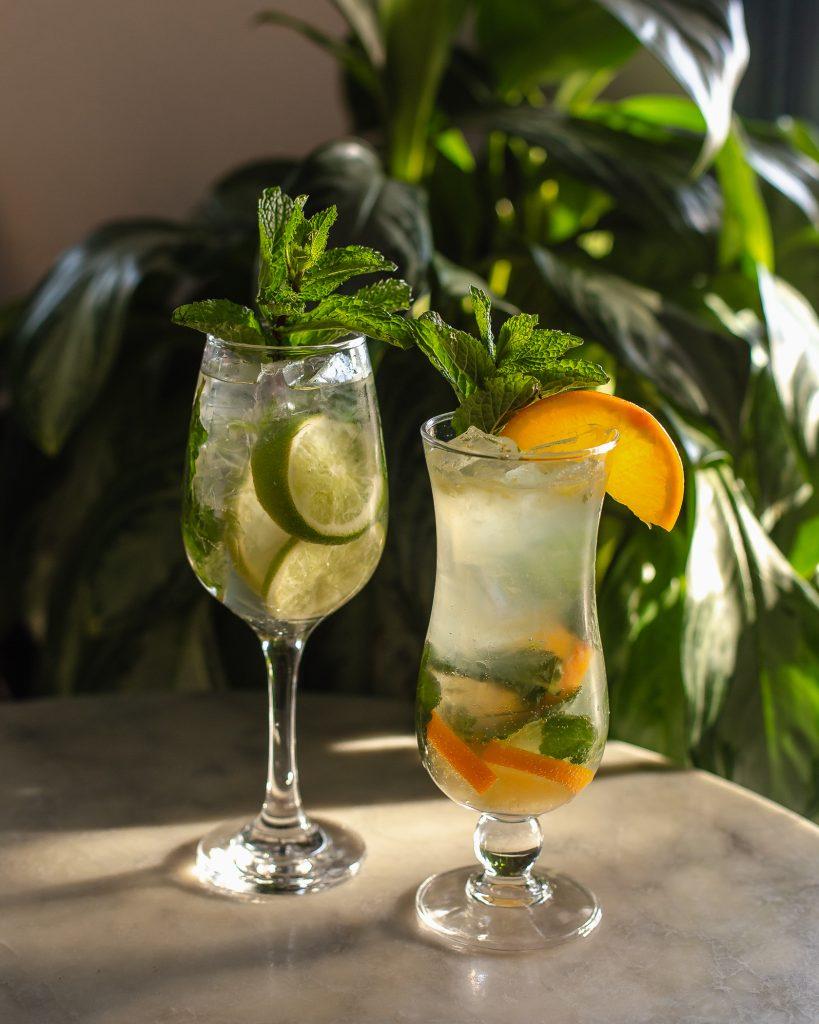 Two mocktails - one pictured in a wine class with limes and a mint garnish. The other in a tall glass with an orange slice